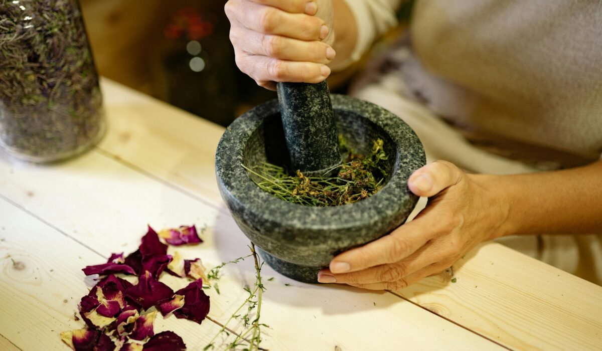 A person grinding herbal ingredients in a stone mortar and pestle with dried petals nearby. — Photo by Yan Krukau on Pexels